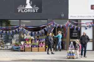 People outside a florist decorated with colorful flowers and Australian flags in a shopping area. A woman at the door, two children with an adult nearby, and a blackboard with store information are visible.