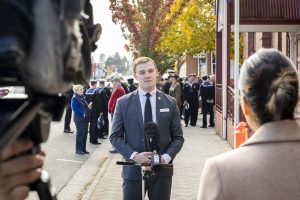 A man wearing a suit speaks into microphones during a street interview. A camera is in the foreground. People in uniform are gathered in the background near a building with red trim, and autumn trees line the street.
