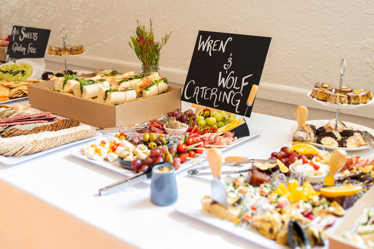 A catered buffet table featuring various foods, including sandwiches, sliced meats, cheeses, olives, grapes, and desserts. A sign reads Wren & Wolf Catering. Decorative flowers are placed near the dishes.