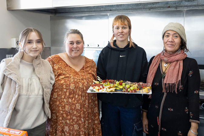 Four people stand in a kitchen. One person holds a platter of assorted foods including fruits and cheese. They are all smiling and dressed casually. The background features a stove and stainless steel surfaces.
