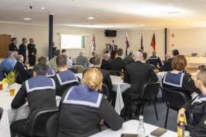 A man in a suit speaks at a podium to an audience of uniformed personnel seated at tables. Flags are displayed behind him. People face forward, listening attentively in a well-lit room.
