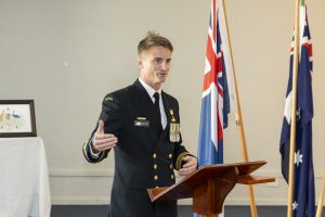 A person in a navy uniform speaks at a wooden podium. They are gesturing with one hand and wearing several medals. Behind them are two flags and a framed certificate on a table.