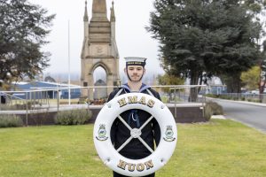 A person in a naval uniform stands on grass holding a large lifebuoy with the text HMAS HUON. A stone monument is visible in the background, surrounded by trees and fog.