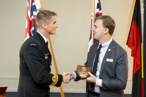 Two men in formal attire shake hands; one wears a naval uniform, and the other a suit with a name badge. Both are holding a wooden plaque with an insignia. Flags are displayed in the background.