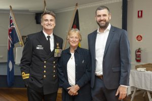 Three people pose together, smiling. On the left is a man in a naval uniform with medals; in the center is an older woman in a dark jacket; on the right is a man in a suit. Flags are visible in the background.