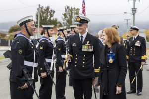A naval officer in uniform, with medals and a sword, converses with a woman wearing a black coat and a blue cross medal. Other uniformed personnel stand in formation in the background, holding rifles. Flags are visible in the overcast setting.