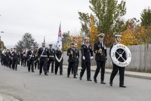 A marching band in navy uniforms parades down a street. One member carries a large bass drum with HMAS Huon written on it. They are followed by others holding instruments and a flag. Trees with autumn leaves line the sidewalk.