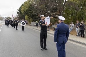 A formal ceremony on a road features uniformed individuals. One person holds a scroll while facing another in uniform. A marching band is seen in the background, and spectators stand along the roadside, capturing the scene on devices. Trees line the street.