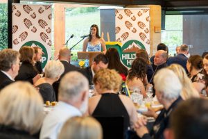 A woman in a blue and white dress speaks at a podium during a formal dinner event. Seated attendees in formal attire listen attentively. HILLTOP HAMP logo is visible on banners behind her.