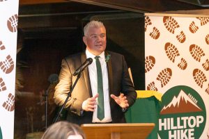 A man in a suit stands at a podium speaking into a microphone. Behind him are banners with brown boot prints and text that reads Hilltop Hike. The setting appears to be an indoor event.