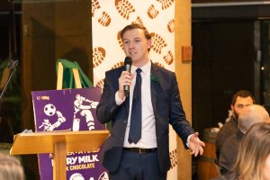 A man in a suit speaks into a microphone at a podium. Behind him, a large purple poster features a soccer player graphic. People are seated at tables, listening. The setting appears to be an event or gathering.