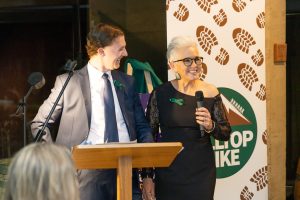 A man in a suit and a woman in a black dress stand at a podium with a microphone. They are smiling. Behind them is a banner with a footprint design and a logo that reads GALLOP F.I.K.E.