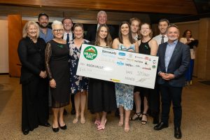A group of smiling people stands indoors holding a large novelty check. All are dressed formally. Logos and the text Hilltop Hike, Pay Huon Valley, and a date are visible on the check.