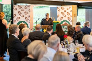 A woman speaks at a podium during an indoor event. Attendees are seated at round tables, listening attentively. Two banners with a logo saying Hike are displayed behind her. The venue has large windows and showcases a view of greenery outside.