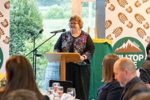 A person standing at a podium, speaking to an audience at a formal event. The backdrop features mountain logos and text. The audience is seated with glasses and plates on the table. The setting appears to be indoors with a view of greenery outside.