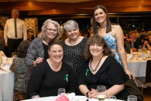 Five people posing together at a formal event. They are dressed in formal attire, with some wearing green ribbons. The background shows tables with guests and decorations. The mood appears cheerful and lively.