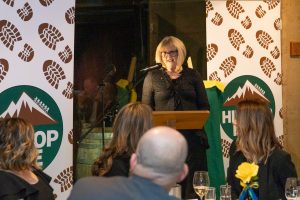 A woman speaks at a podium in front of an audience. Behind her are banners with hiking boot prints and a mountain logo. People in the audience are seated at tables, with wine glasses and a vase containing a yellow rose.