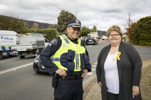 A police officer in a high-visibility vest and a woman wearing a yellow ribbon stand smiling on a street. Police cars and a roadblock are visible in the background. It is a cloudy day with trees lining the road.