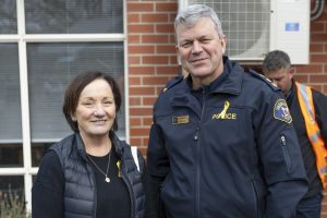 A man in a police uniform and a woman in a black vest smile outdoors. They both wear yellow ribbon pins. A man in an orange jacket stands in the background near a brick building with windows and an air conditioning unit.