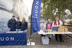A group of people at an outdoor event stand behind tables with baked goods and snacks. A flag with the text Oceans Ahead. Always. is displayed. A mural and trees are in the background.