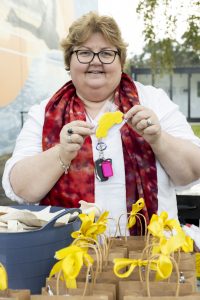 A person with glasses and a red scarf holds up a yellow car-shaped cookie. In front of them are bags with yellow ribbon bows. A mural is partially visible in the background. It appears to be an outdoor setting.