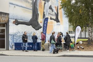 A group of people chatting near a table with promotional banners and a small tent outdoors. A large mural of flying geese is on the building behind them. A black dog stands near the group. Trees and a sidewalk are nearby.
