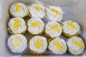 A dozen vanilla cupcakes with white frosting, each topped with a yellow ribbon decoration. The cupcakes are placed in a clear plastic container.
