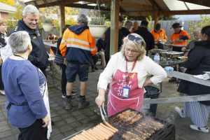 A woman wearing glasses and a red apron cooks sausages and skewers on a barbecue. People stand and sit around under a gazebo, some in orange work gear. Potted plants hang overhead. The atmosphere appears relaxed and social.