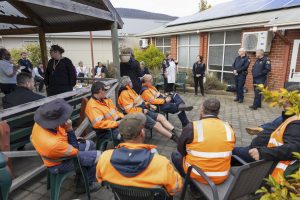 A group of people, some in orange safety vests, gather outdoors in a courtyard. They are seated and standing, listening to a few speakers by a brick building. The area is partially covered by a wooden canopy.