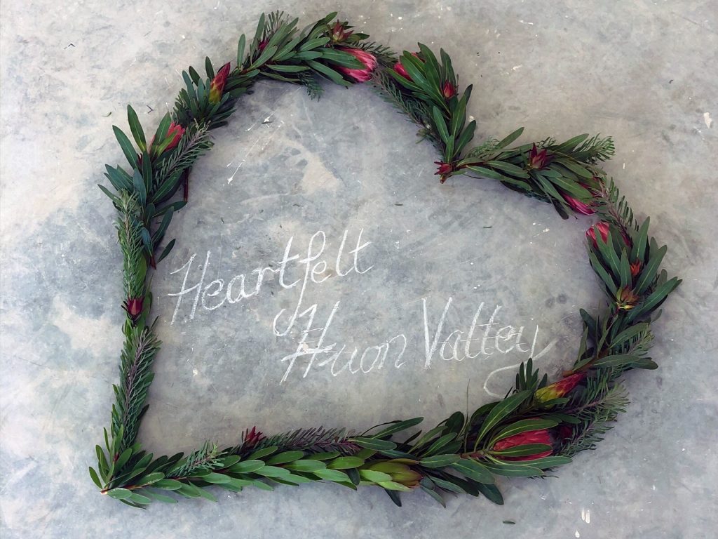 A heart-shaped wreath made of green leaves and red flowers lies on a stone surface. Inside the heart, the words Heartfelt Huon Valley are written in white chalk.