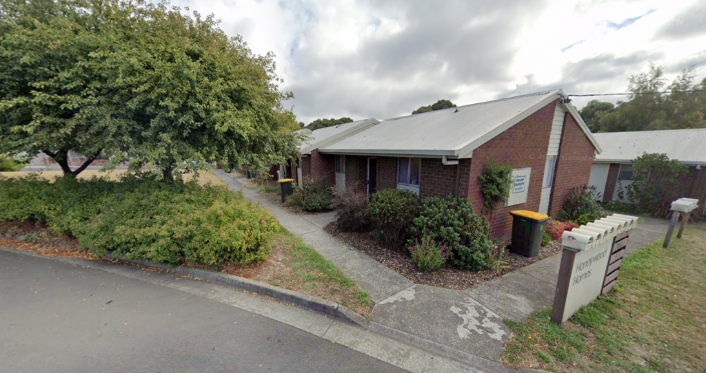 A single-story brick building with a metal roof, surrounded by shrubs and trees, with pathways leading to entrances and a yellow-lidded bin near the doorway. A sign reading Doctors Office is visible in front.