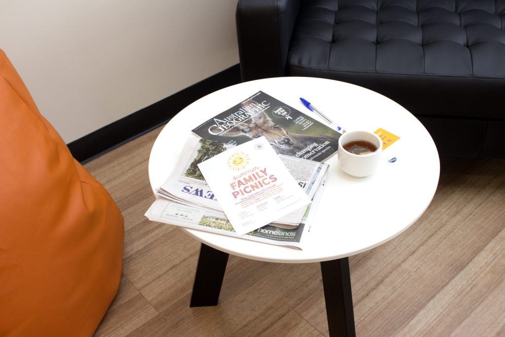 A white round table with a cup of coffee, a blue pen, and various magazines, including one titled Family Picnics and another Australian Geographic. An orange cushion and black leather sofa are in the background.
