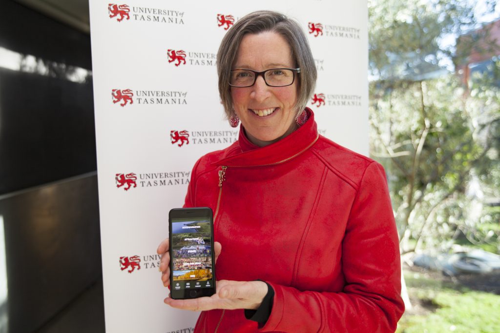 A person in a red jacket stands in front of a University of Tasmania backdrop, smiling and holding a smartphone displaying an app with landscape images.
