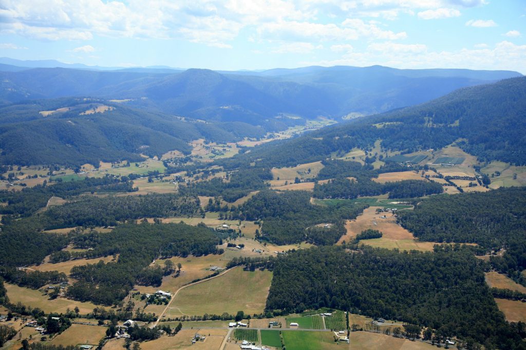 Aerial view of a lush valley surrounded by mountains. The landscape features patches of forests, farmland, and scattered houses. The sky is partly cloudy and bright, adding depth to the rolling terrain.