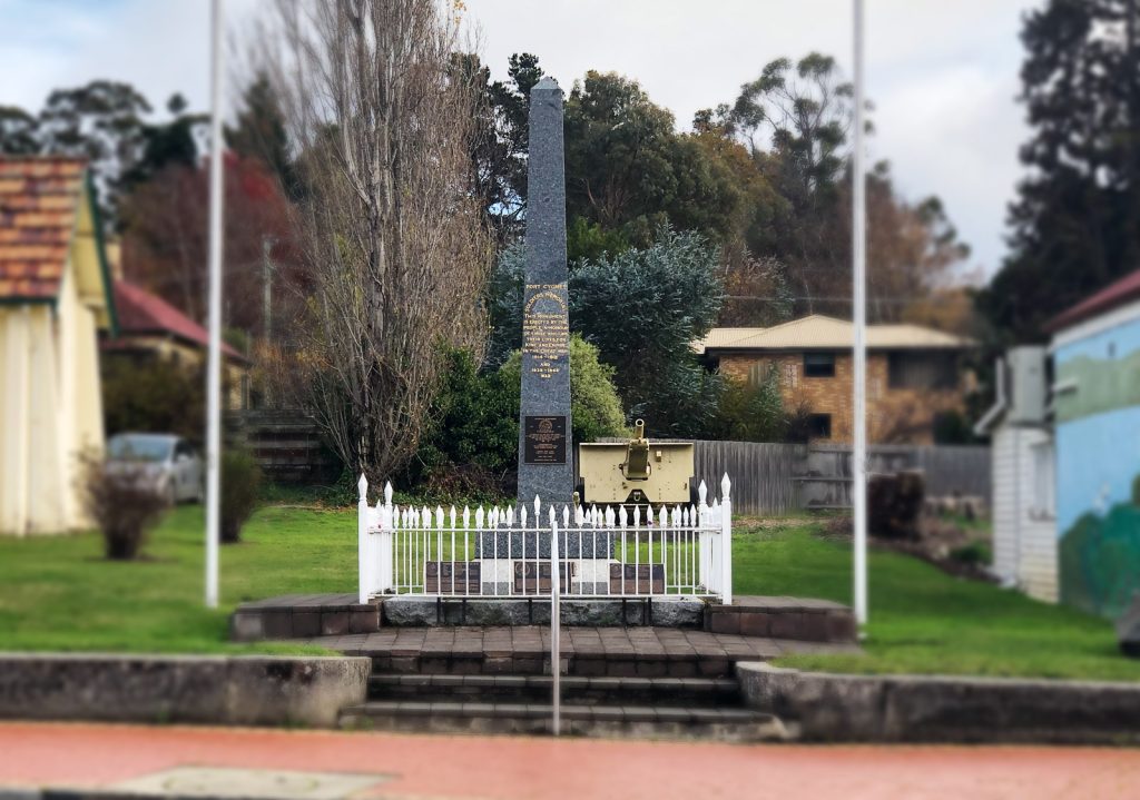 A tall stone war memorial stands in a small fenced area with plaques on its surface. It is surrounded by greenery and flanked by flagpoles. Two buildings are visible in the background. The sky is overcast.
