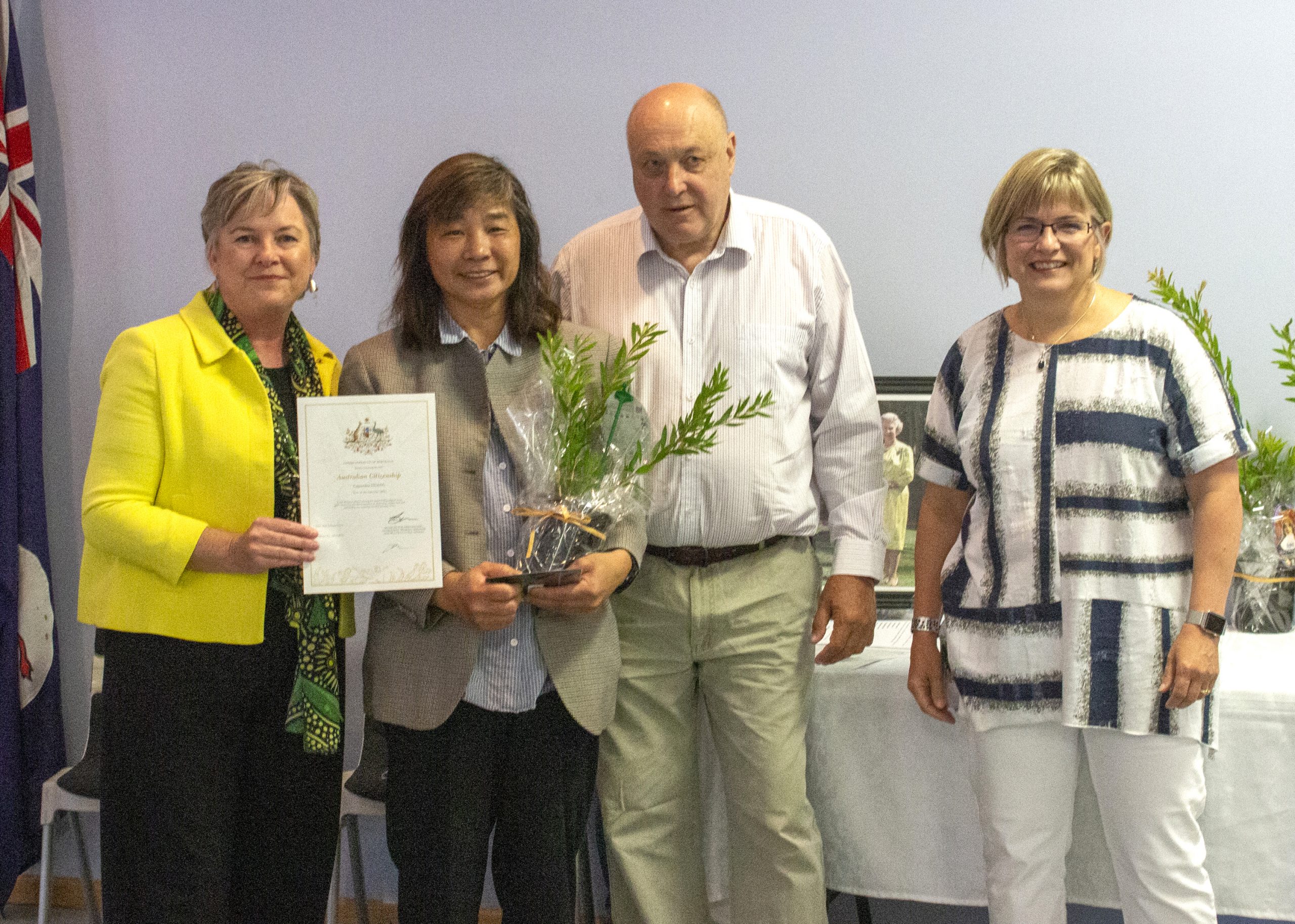 Four people are standing together in a room. One person is holding a certificate, and another is holding a plant wrapped in transparent paper. They are dressed in formal and semi-casual attire, with an Australian flag visible in the background.