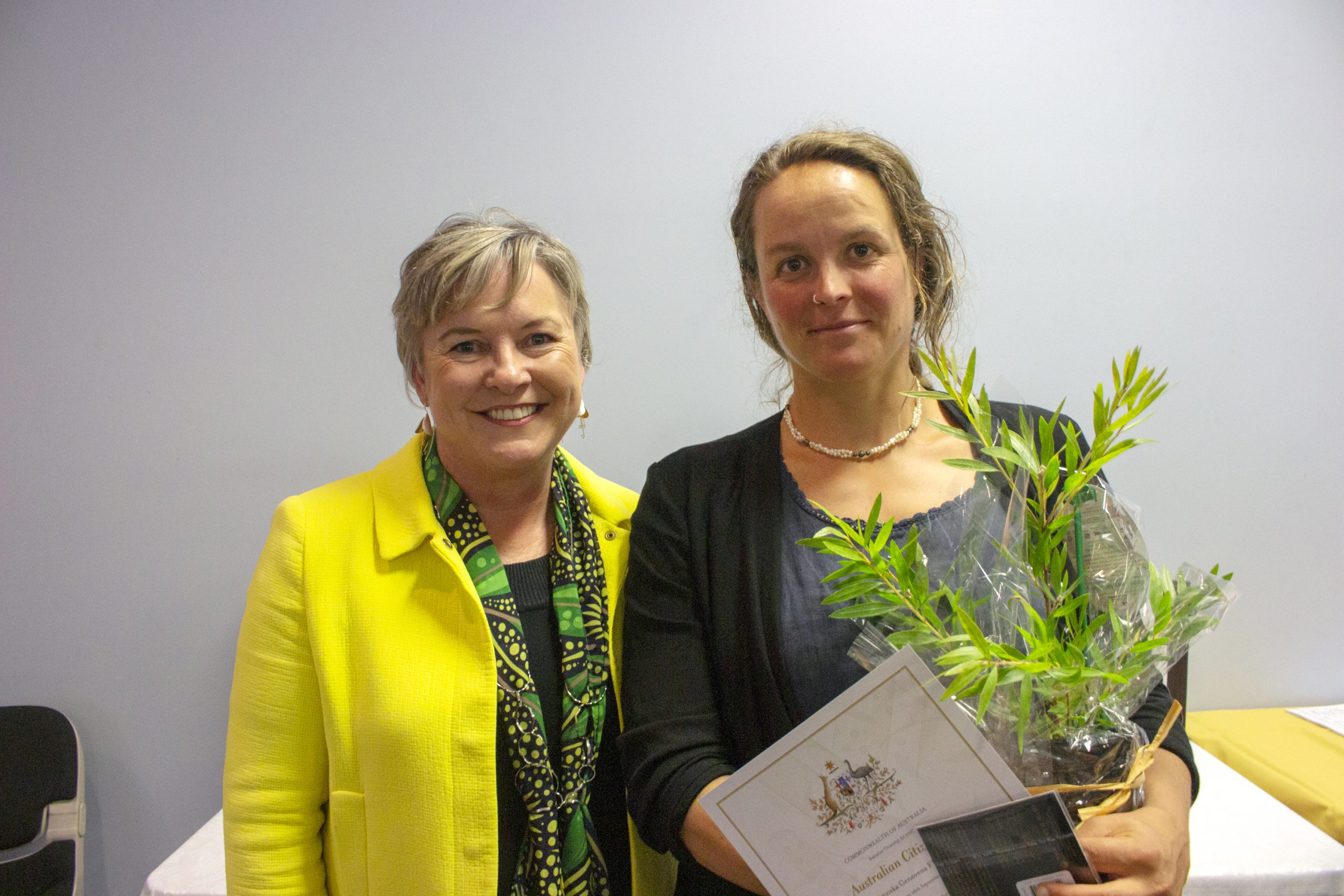 Two women stand side by side, smiling. The woman on the left is wearing a bright yellow jacket. The woman on the right holds a small plant and a certificate. They appear to be at an event or ceremony indoors.