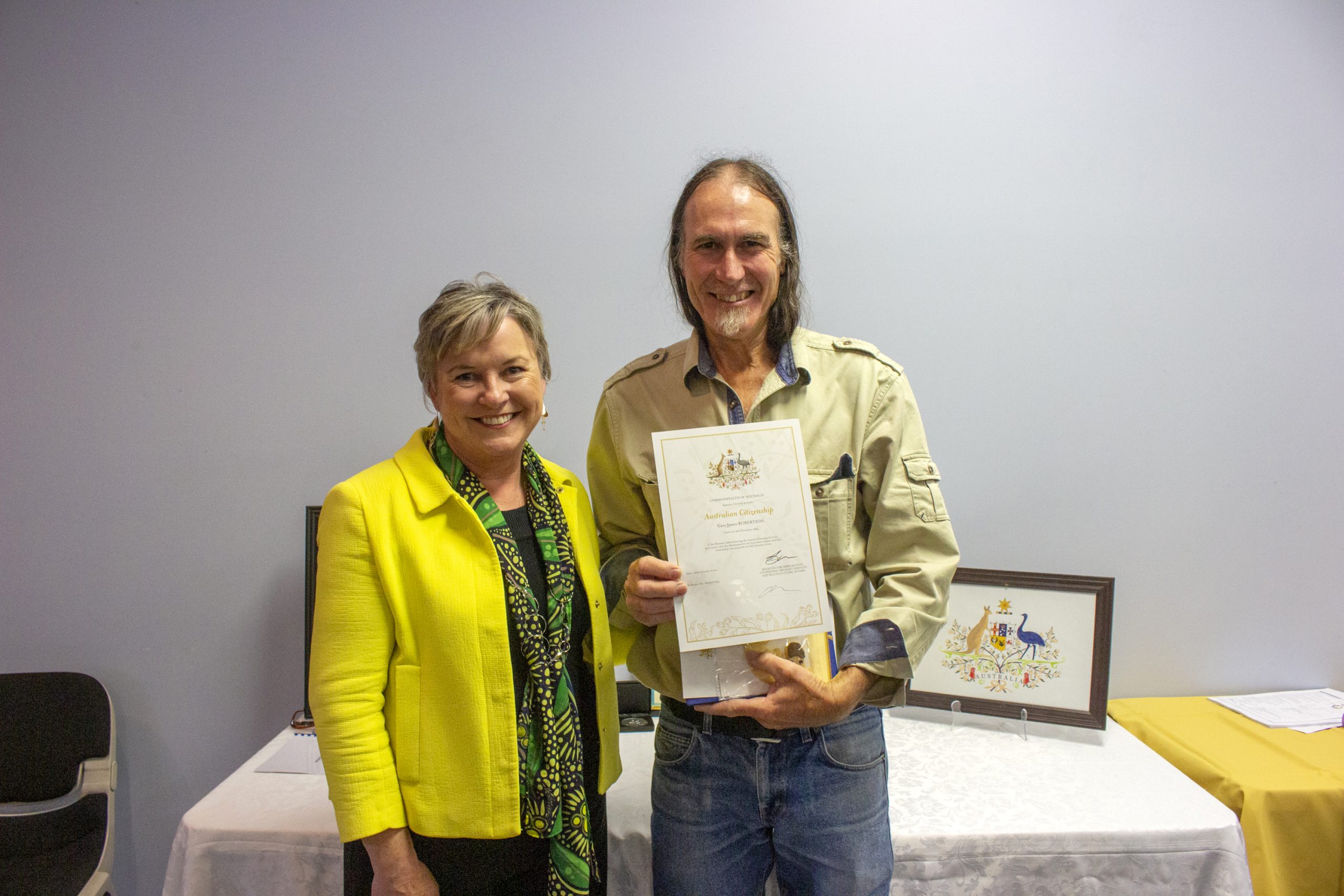 Two people standing indoors, smiling. The person on the right is holding a certificate. A table with a framed document and a yellow cloth is in the background. The person on the left is wearing a bright yellow jacket.