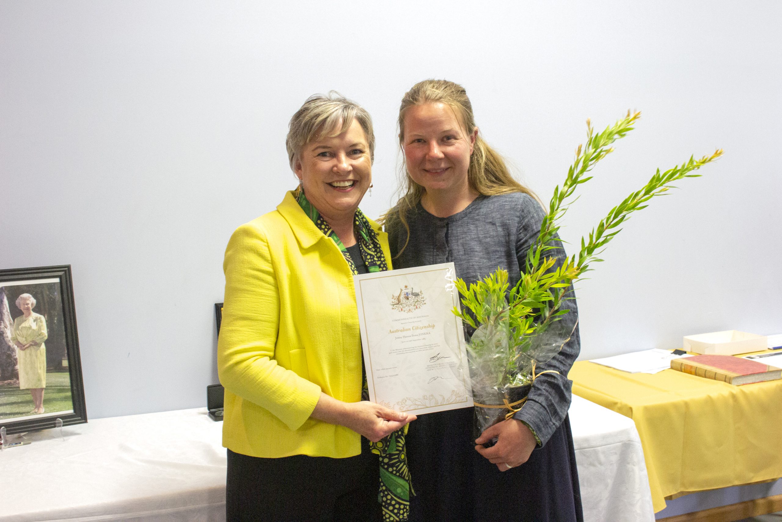 Two women stand together smiling. The woman on the left, in a yellow jacket, is holding a certificate. The woman on the right, in a dark outfit, holds a small plant. A framed photo and a yellow cloth are on a table in the background.