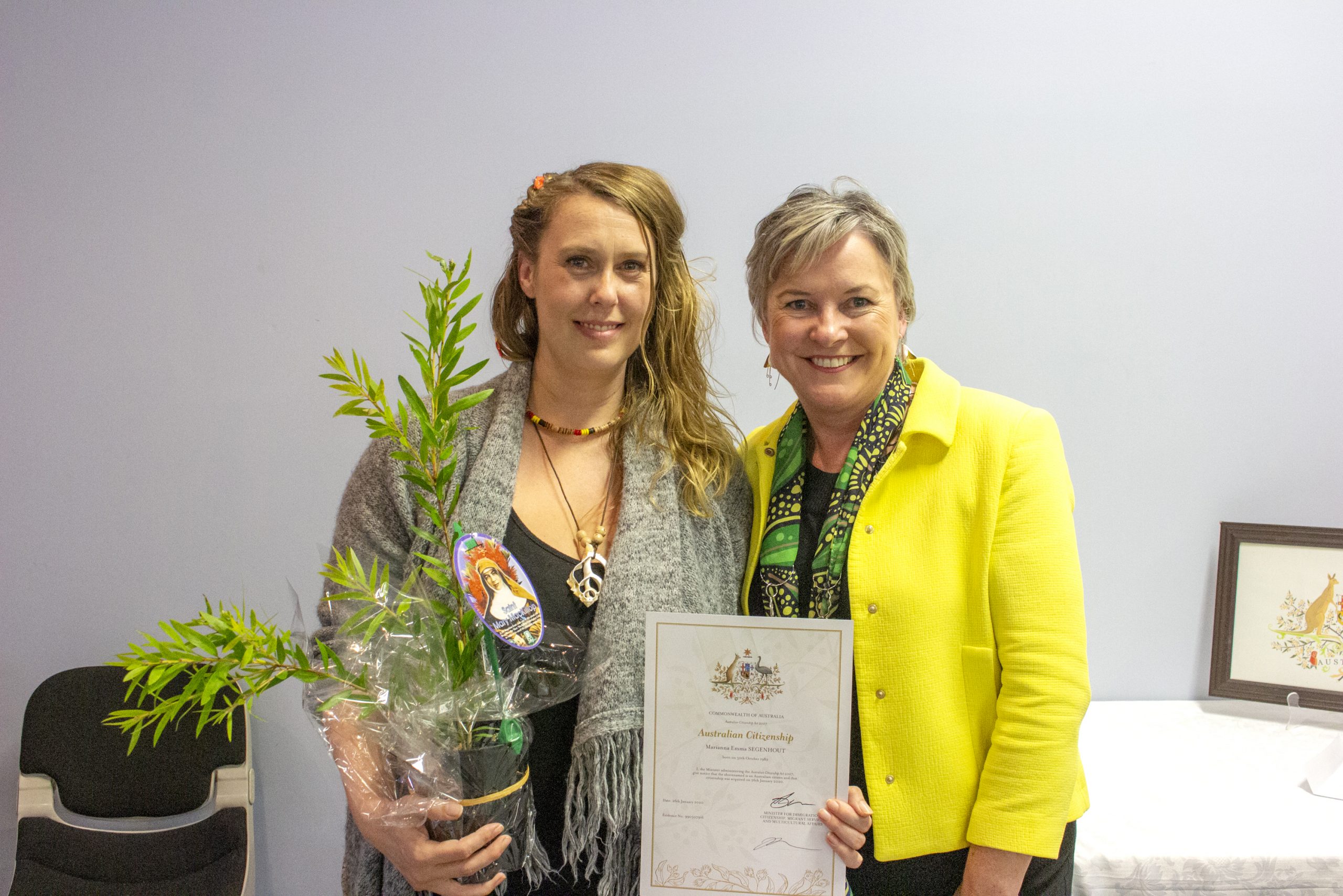 Two women stand together, one holding a potted plant and the other a certificate. Both are smiling. The woman on the left has long hair and wears a necklace, while the woman on the right is in a yellow jacket. A small table with papers is in the background.