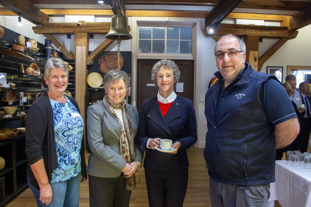 Four people stand indoors, smiling at the camera. The setting is a warmly lit room with wooden beams and shelves displaying pottery. One person holds a cup and saucer. They are casually dressed, and a few others are visible in the background.