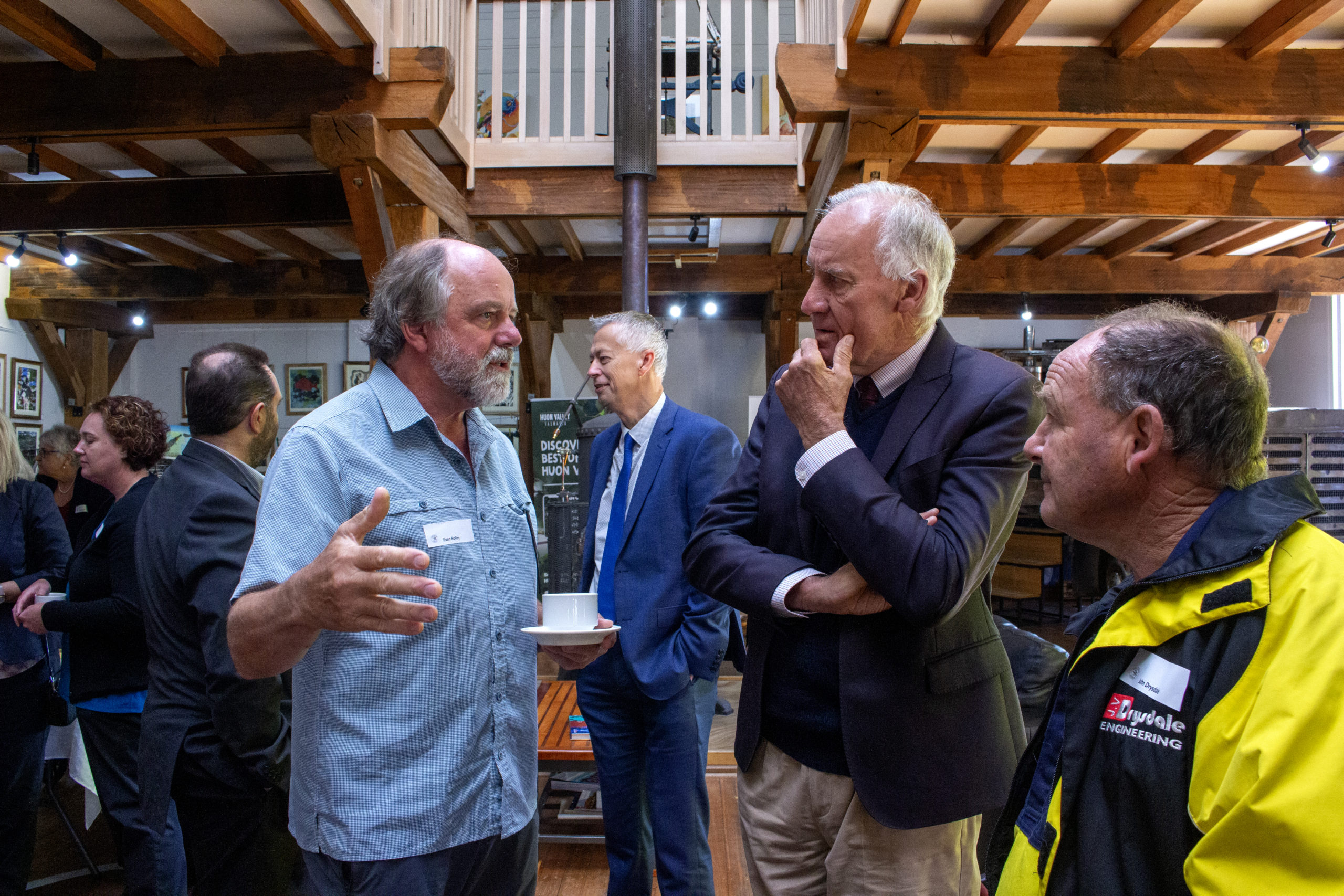 Four men are engaged in conversation in a wooden-beamed room with art on the walls. One man gestures with his hands, while two others listen intently. The fourth man wears a yellow jacket with a logo. Other people are in the background.