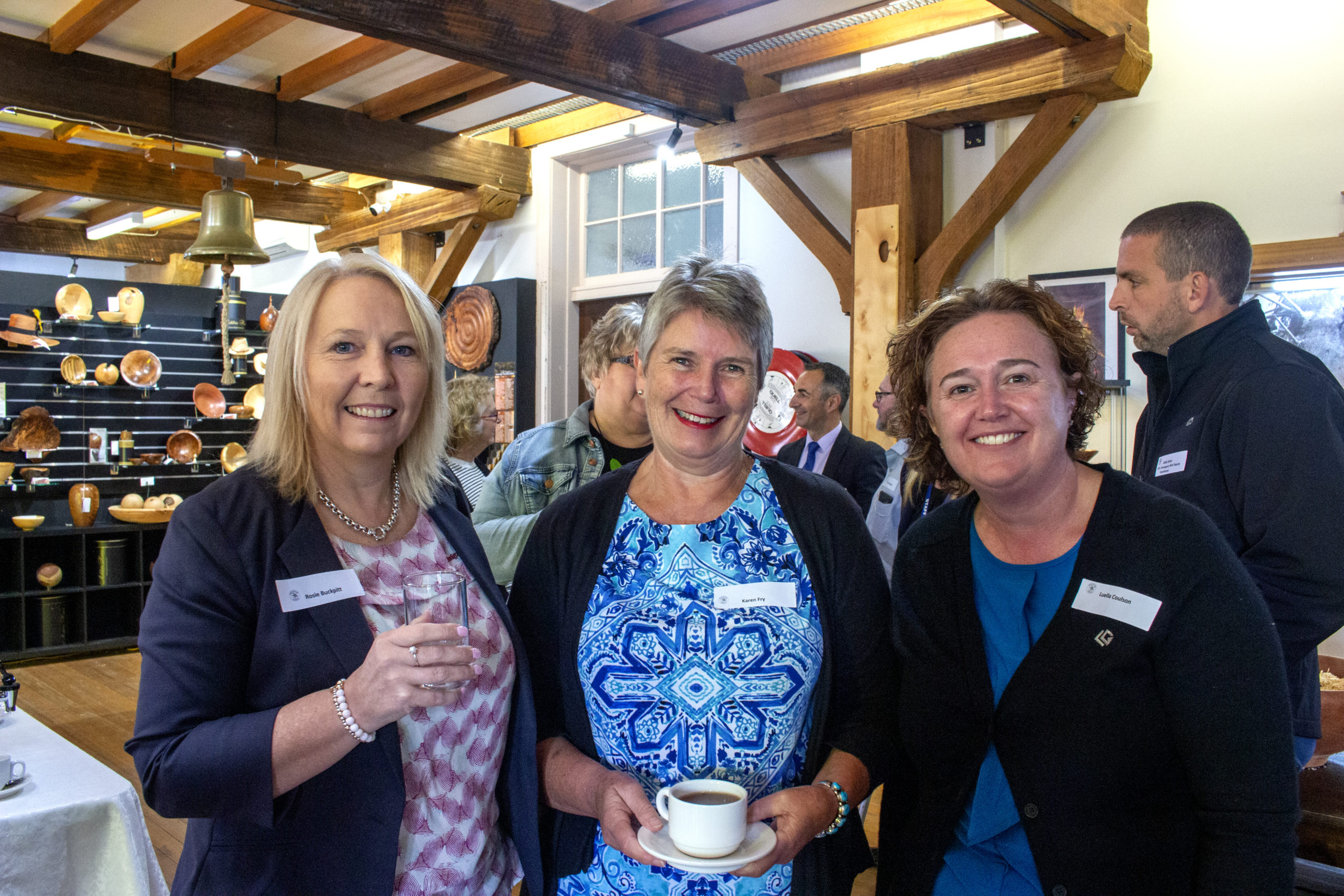 Three women smiling and posing for a photo indoors. One woman holds a coffee cup. They wear name tags and formal attire. In the background, there are wooden sculptures and other people engaged in conversation. The room has exposed wooden beams.