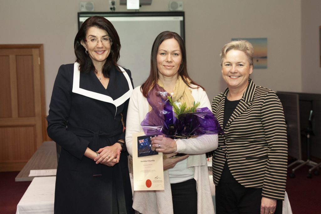 Three women are standing indoors, smiling at the camera. The woman in the middle holds a certificate and a bouquet with purple wrapping. They are dressed in business attire, and a door and a whiteboard are visible in the background.