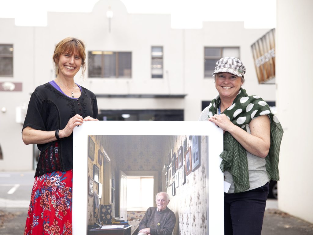 Two women are standing outdoors, smiling and holding a large framed photograph. The photograph depicts an older person sitting in a vintage-styled room with patterned walls and framed pictures. .