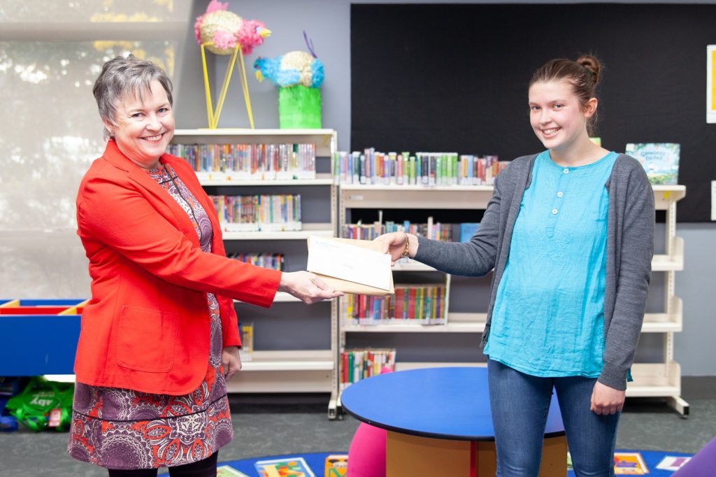 A woman in a red blazer hands an envelope to a young woman in a blue shirt and gray cardigan. They are standing in a room with bookshelves and a round table, smiling.