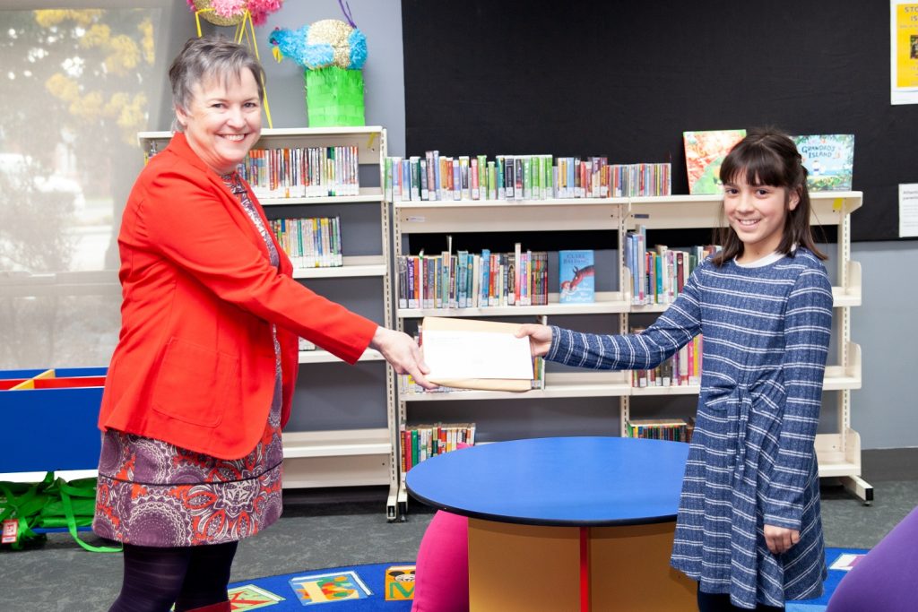 A woman in a red jacket and patterned dress hands an envelope to a smiling girl in a striped dress. They are standing in a room with bookshelves filled with colorful books. There is a round table with bright chairs in the foreground.