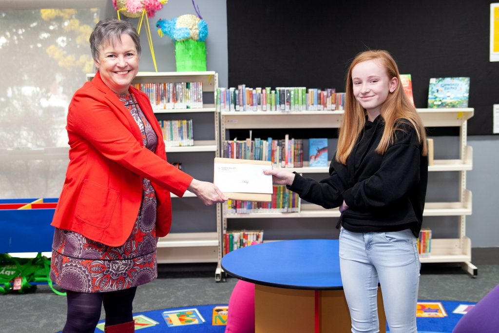 A woman in a red blazer hands an envelope to a young girl with long red hair in a library setting. Bookshelves filled with colorful books are in the background, and they stand near a round blue and yellow table.