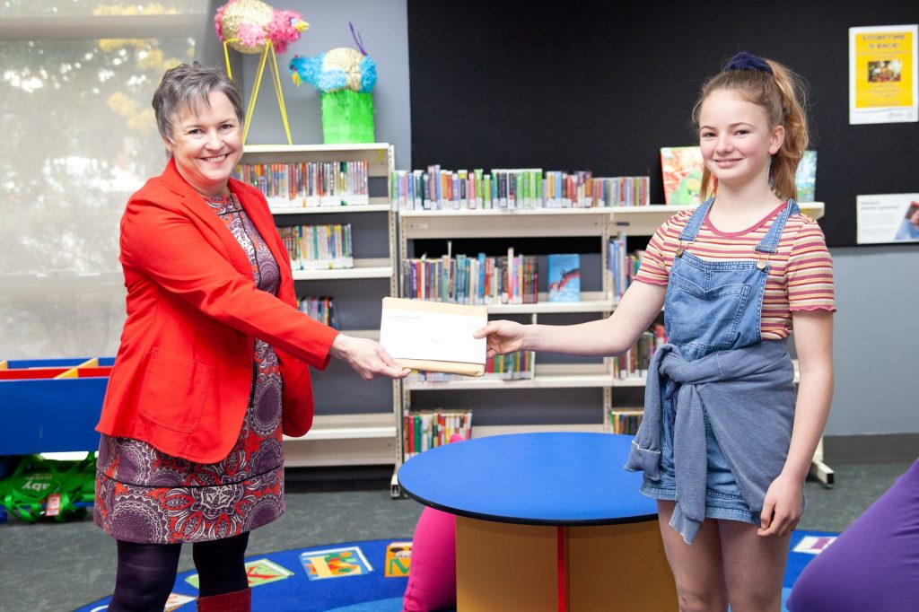 A woman in a red jacket hands a certificate to a smiling girl in denim overalls. They are in a library with colorful decorations and bookshelves.