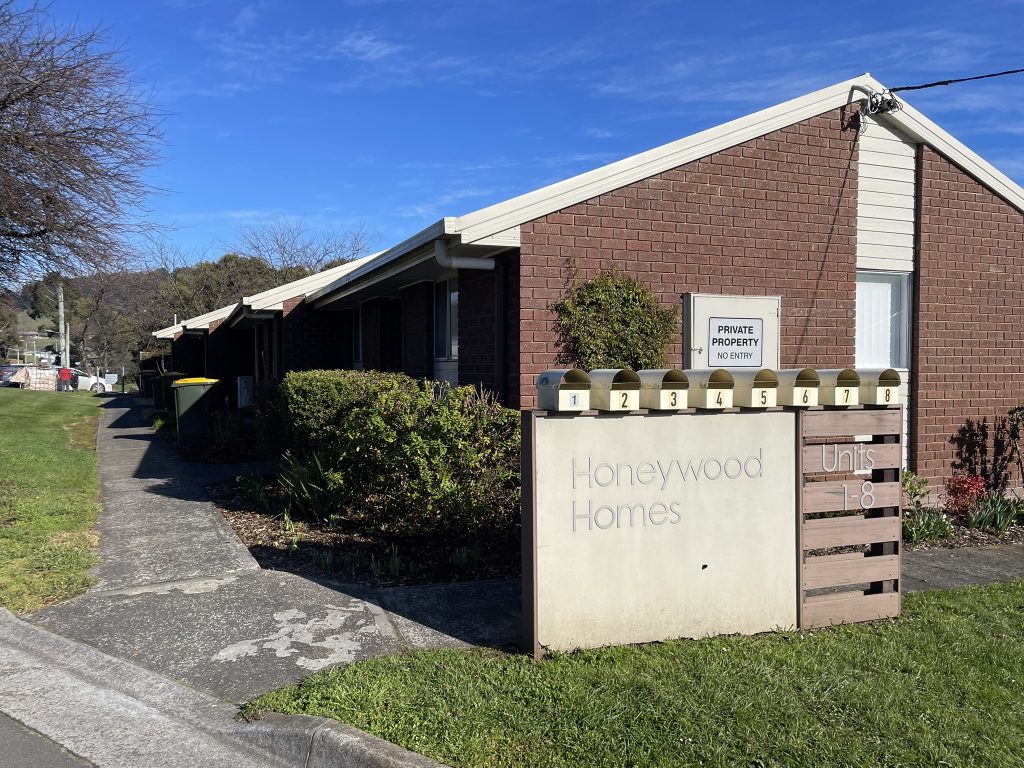 A row of brick units at Honeywood Homes with eight yellow mailboxes labeled 1–8 on a concrete sign at the entrance. A Private Property No Entry sign sits on top. Shrubs and a sidewalk are visible under a clear blue sky.